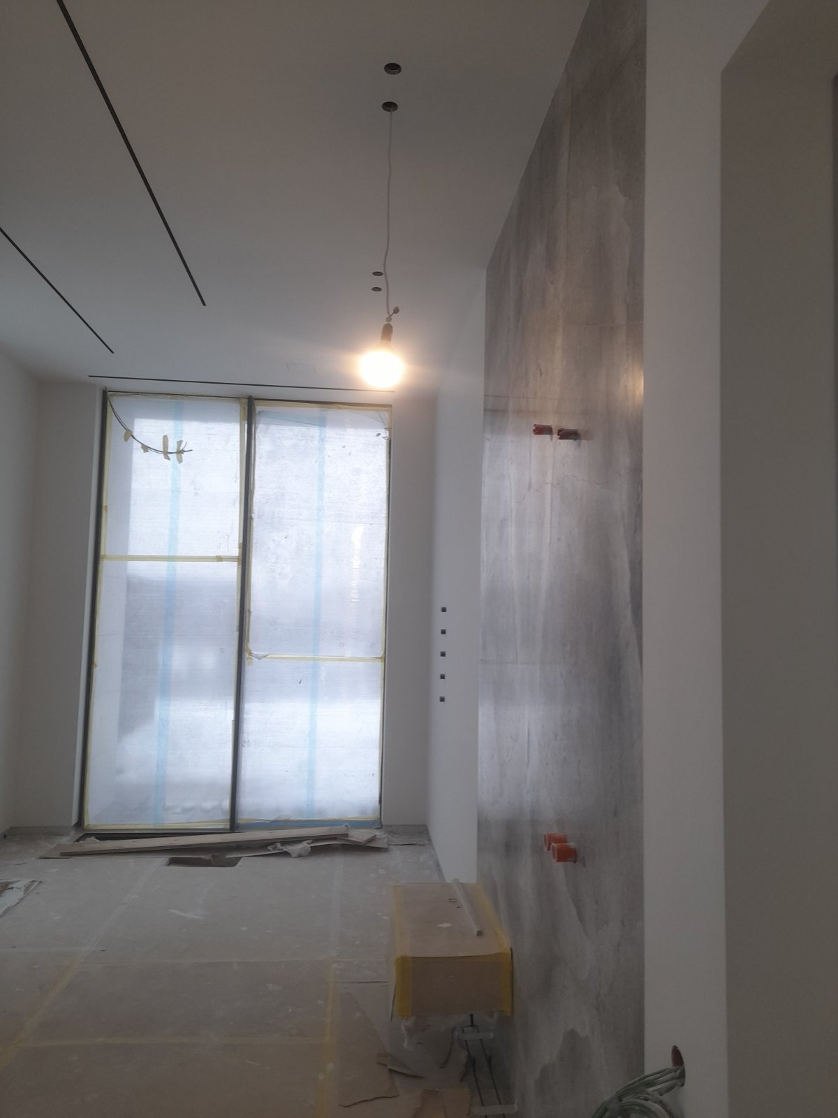 Wnętrze w trakcie wykańczania: ściana z efektem betonu, zabezpieczone okno folią, widoczne punkty elektryczne i lampa wisząca. Minimalistyczny design w szarościach.