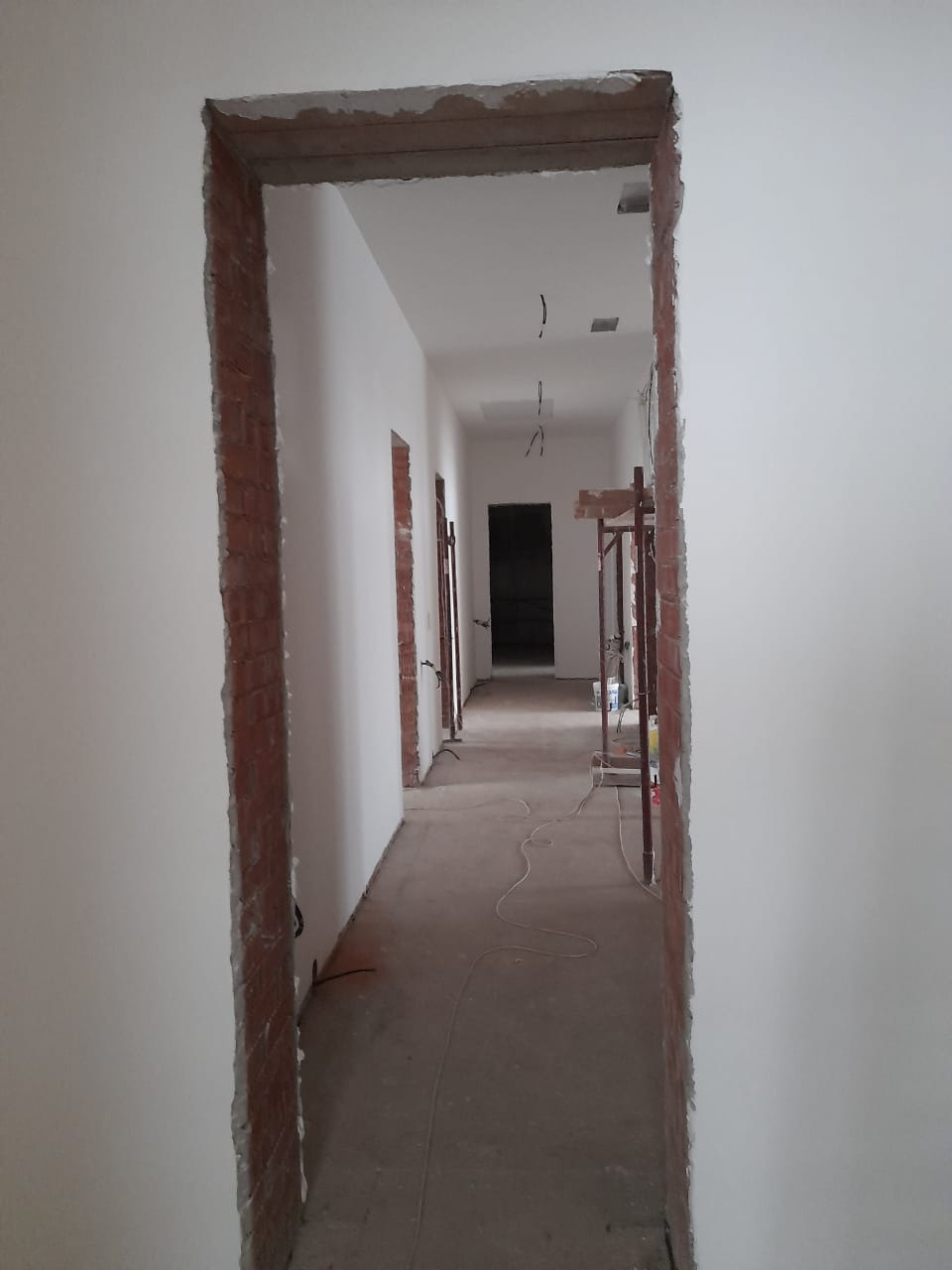 Korytarz w trakcie remontu: surowe ściany, widoczne cegły wokół ościeżnic, w tle rusztowanie. Etap przygotowania do wykończenia wnętrz.