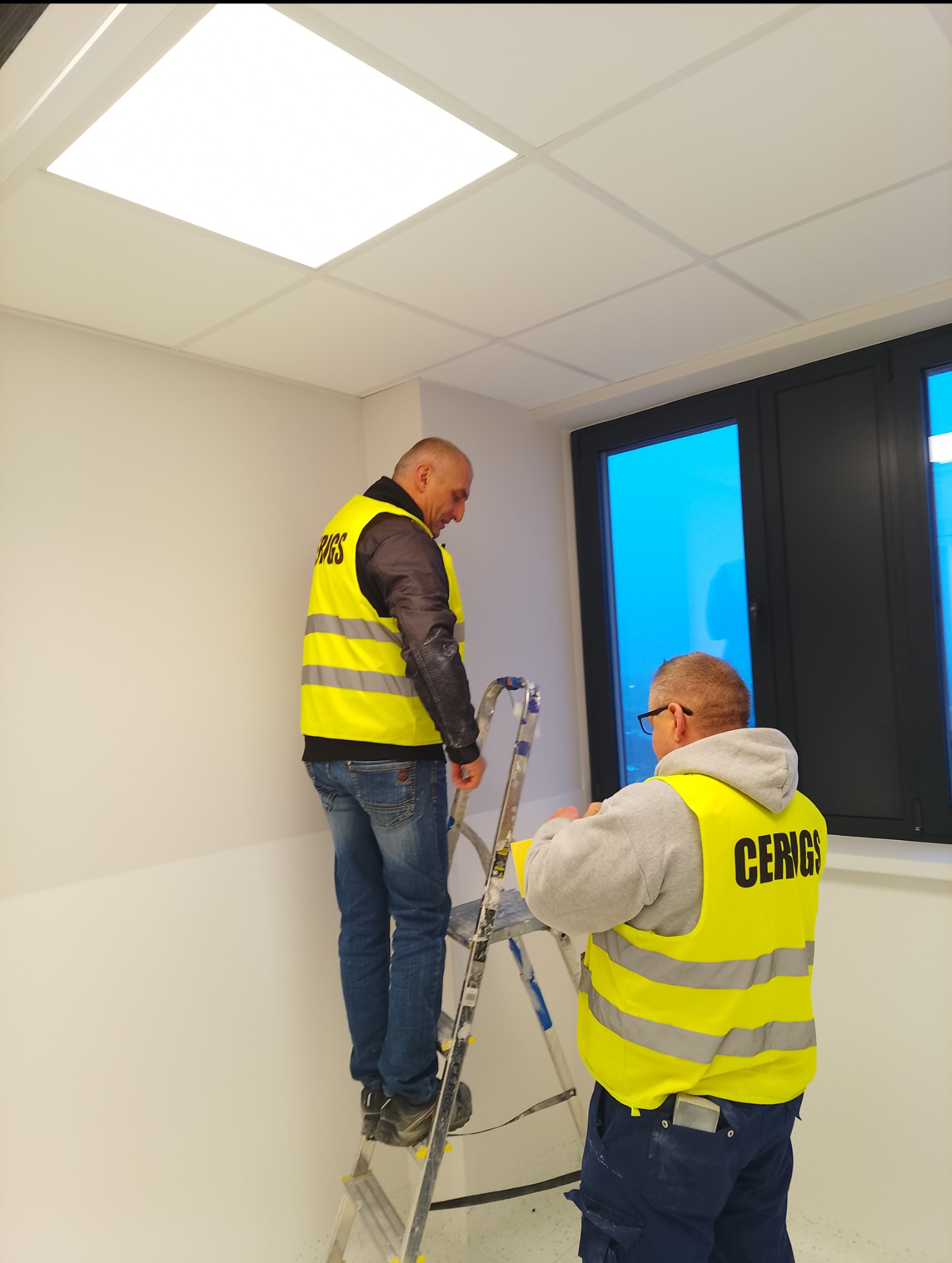 Dwóch pracowników w kamizelkach na drabinie montuje kasetonowe oświetlenie sufitowe w jasnym pomieszczeniu z oknami. Widok od dołu, skupienie na suficie i pracownikach.