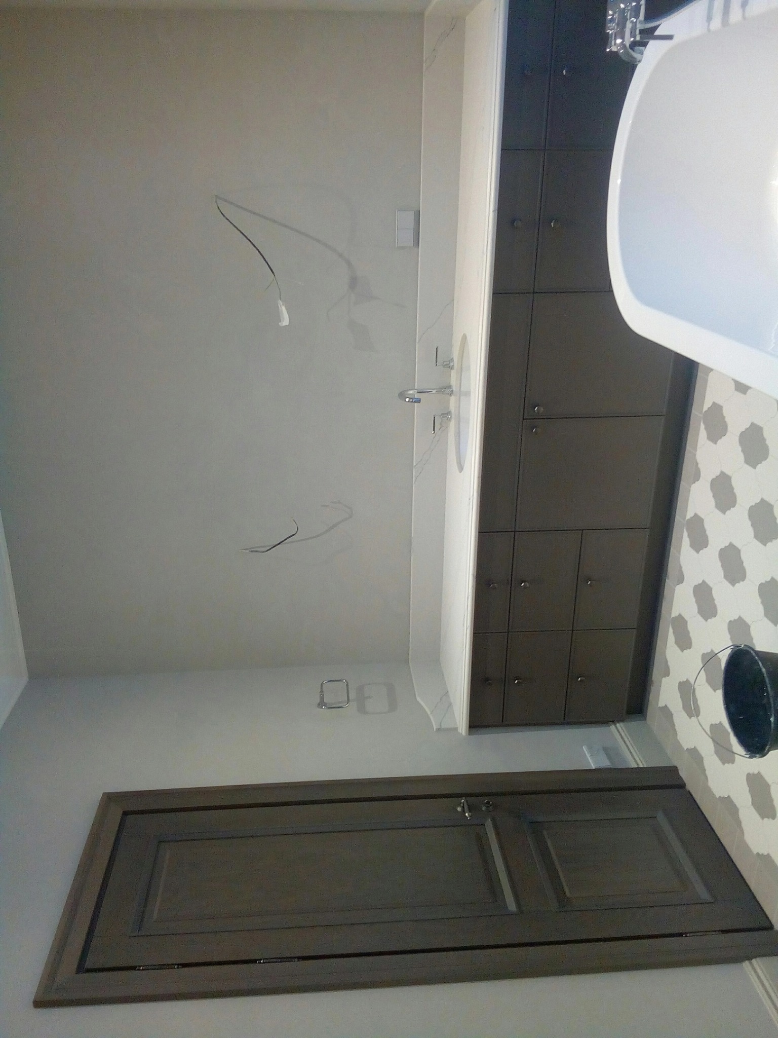 Wykończona łazienka z ciemnymi szafkami, białą umywalką i wanną. Na podłodze wzorzyste płytki. Widoczne wystające kable ze ściany. Ujęcie z dołu.