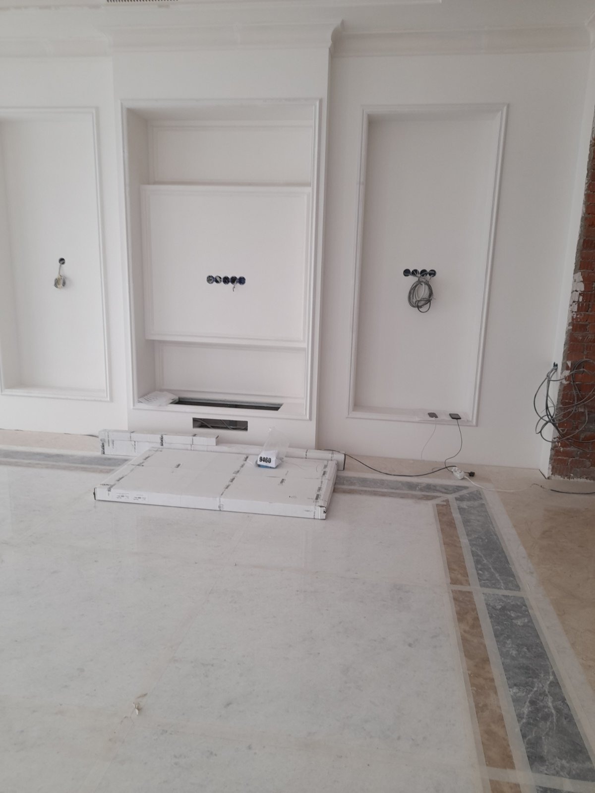 Białe ściany z dekoracyjnymi wnękami i listwami, widoczne gniazdka elektryczne, podłoga z marmurowym wzorem, kartony na podłodze. Wnętrze w trakcie wykończenia.