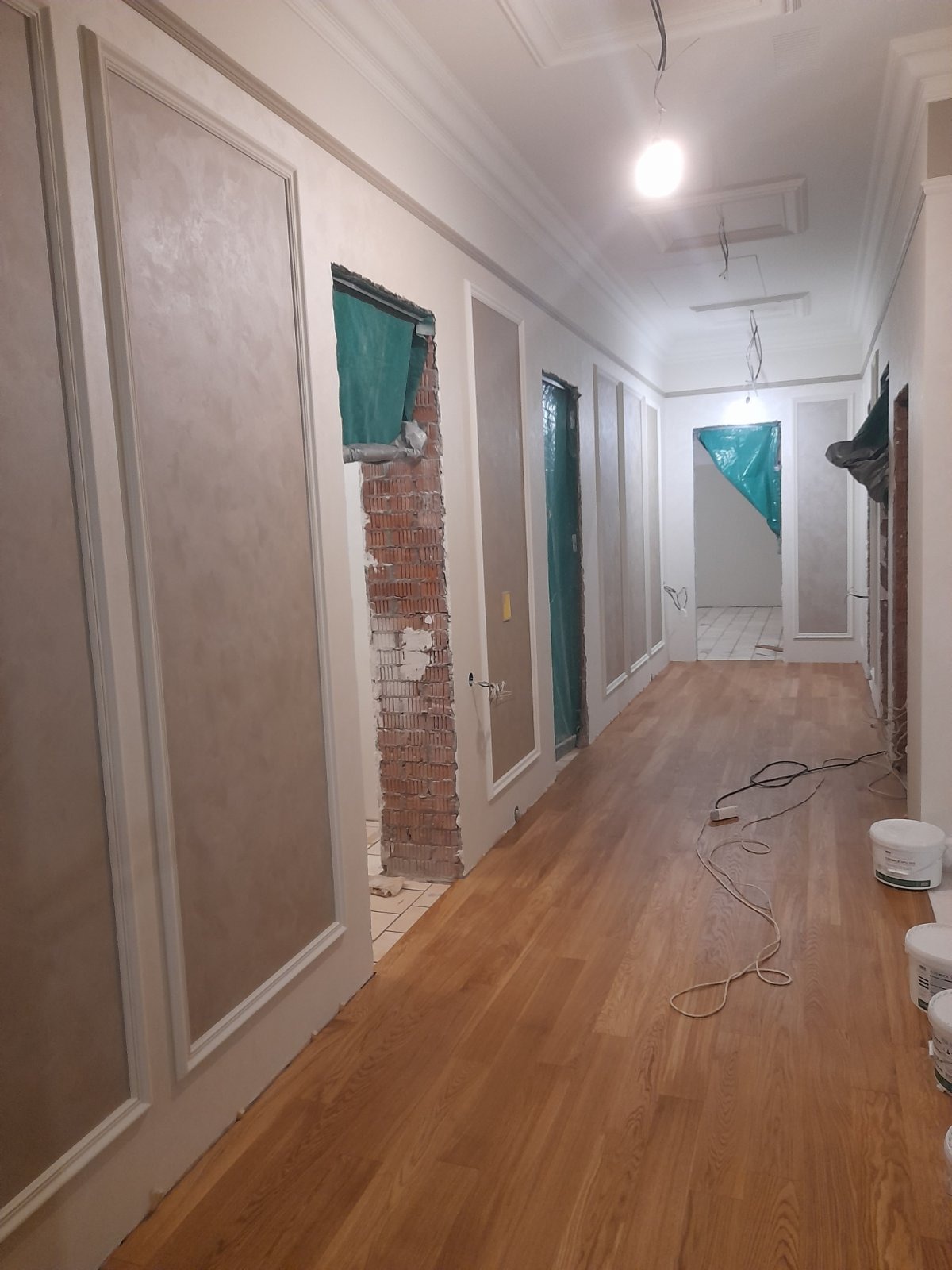Korytarz w trakcie prac wykończeniowych: widoczne panele na ścianach, odsłonięta cegła wokół ościeżnic, drewniana podłoga i kable. Etap prac w jasnym wnętrzu.