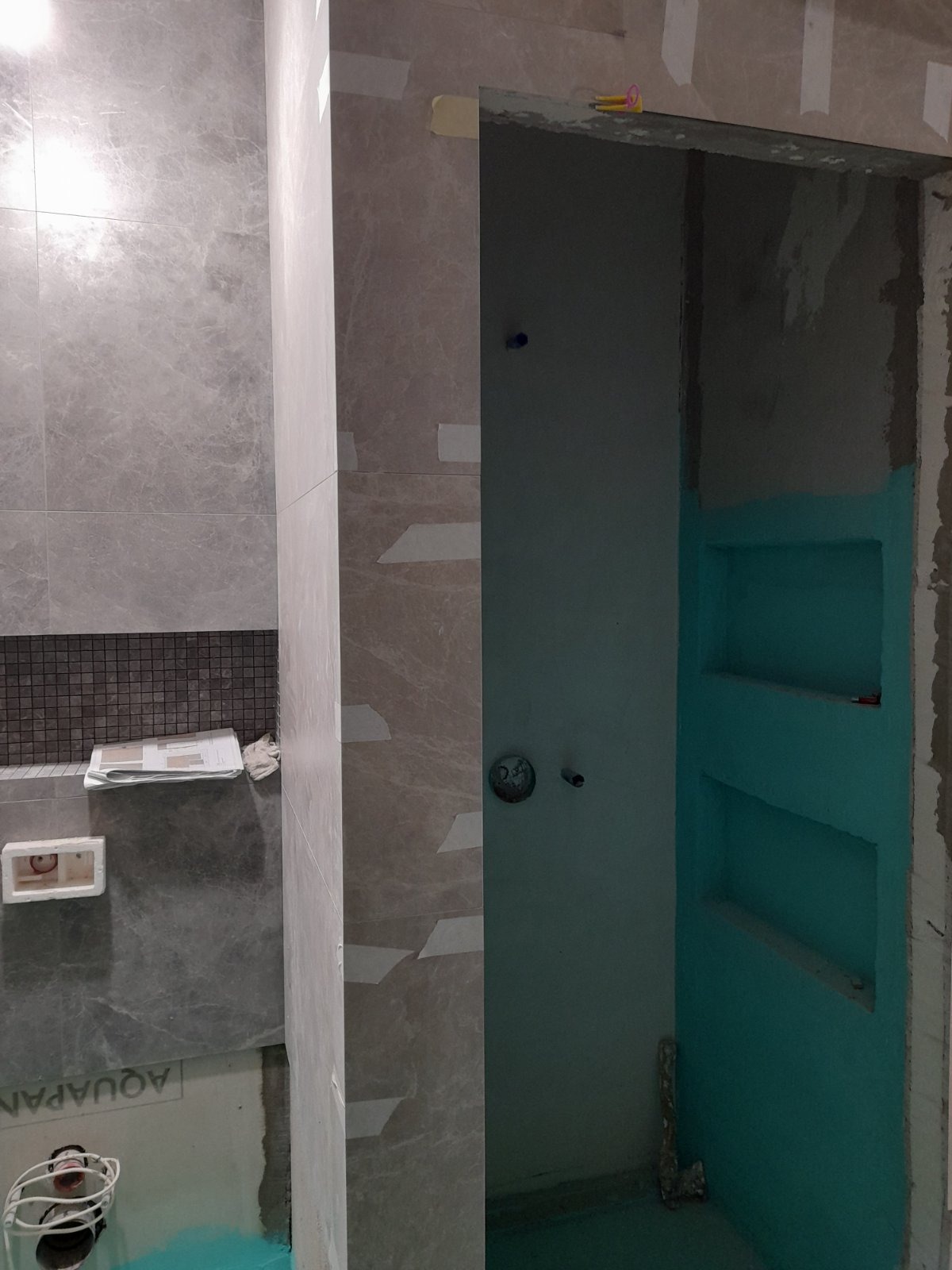Wnętrze łazienki w trakcie wykańczania: szare płytki imitujące marmur, hydroizolacja wnęki prysznicowej w kolorze turkusowym, widoczne materiały budowlane i narzędzia.