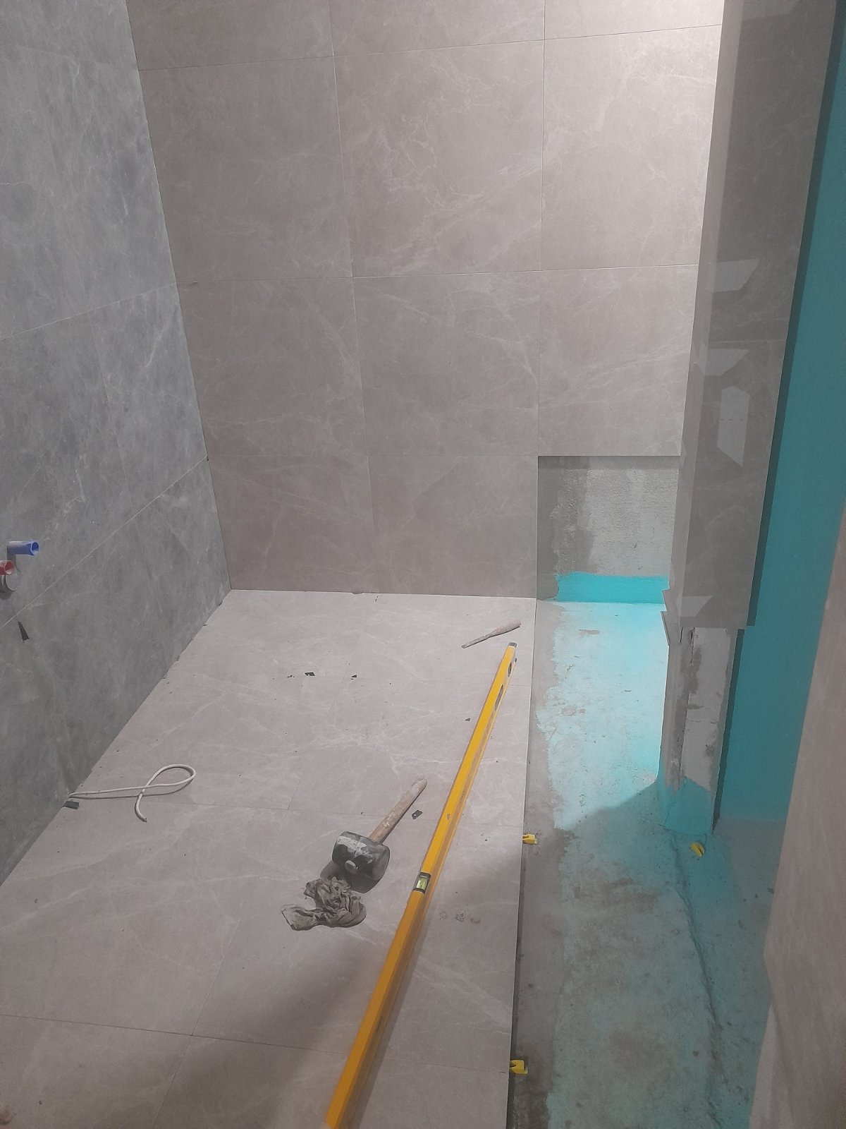 Układanie płytek w łazience: szare płytki na ścianach i podłodze, poziomica, gumowy młotek. Widoczna hydroizolacja w strefie prysznica. Prace glazurnicze w toku.