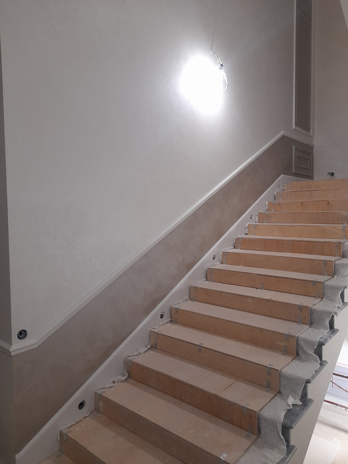 Schody w trakcie wykończenia z zamontowanymi płytami. Widoczne przygotowane ściany i oświetlenie. Detal wykończenia wnętrza w jasnych barwach.