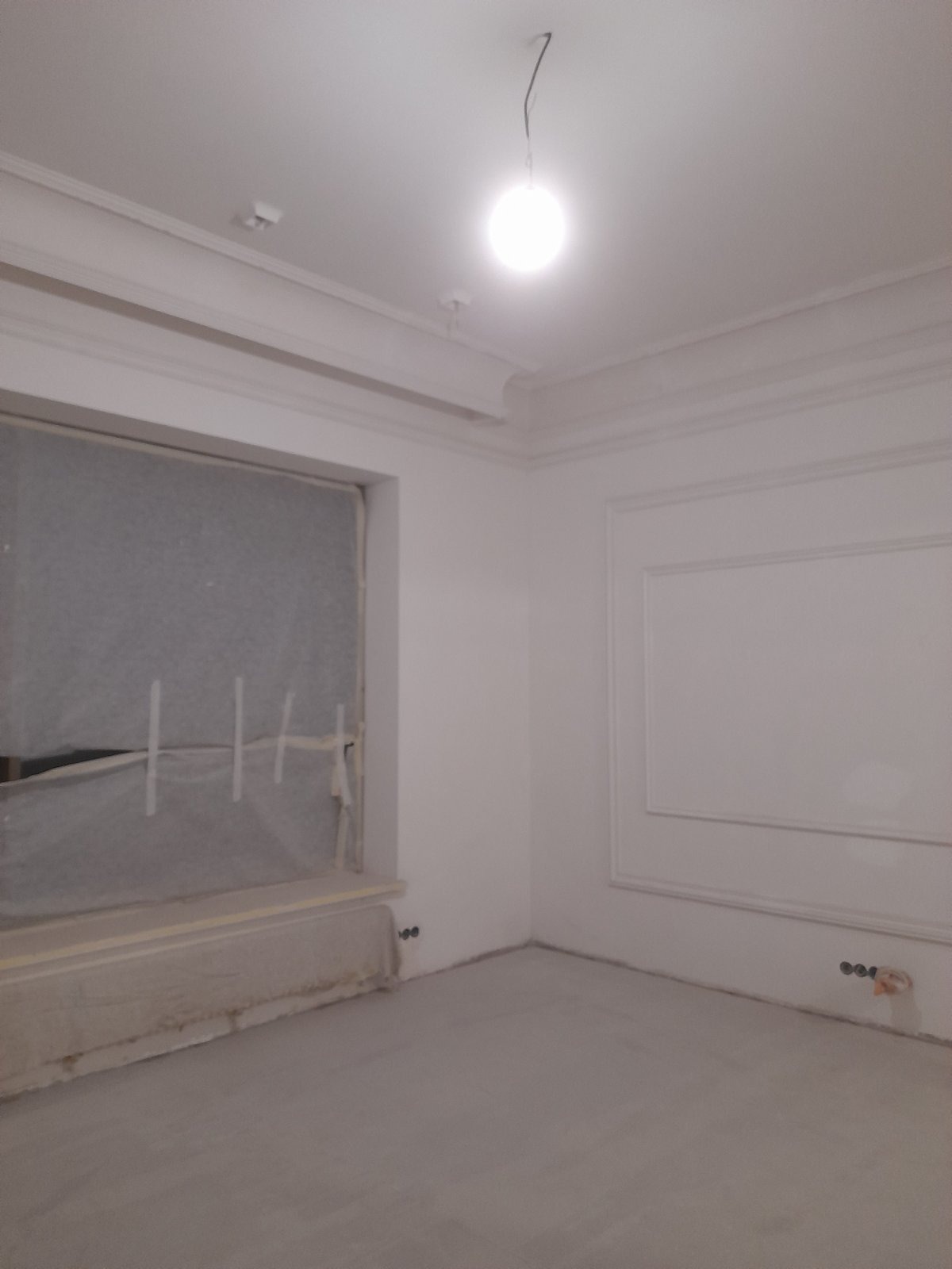 Białe wnętrze w trakcie wykańczania: okno zabezpieczone folią, ściany z dekoracyjnymi listwami, surowa podłoga, widoczne gniazdka elektryczne. Oświetlenie sufitowe.