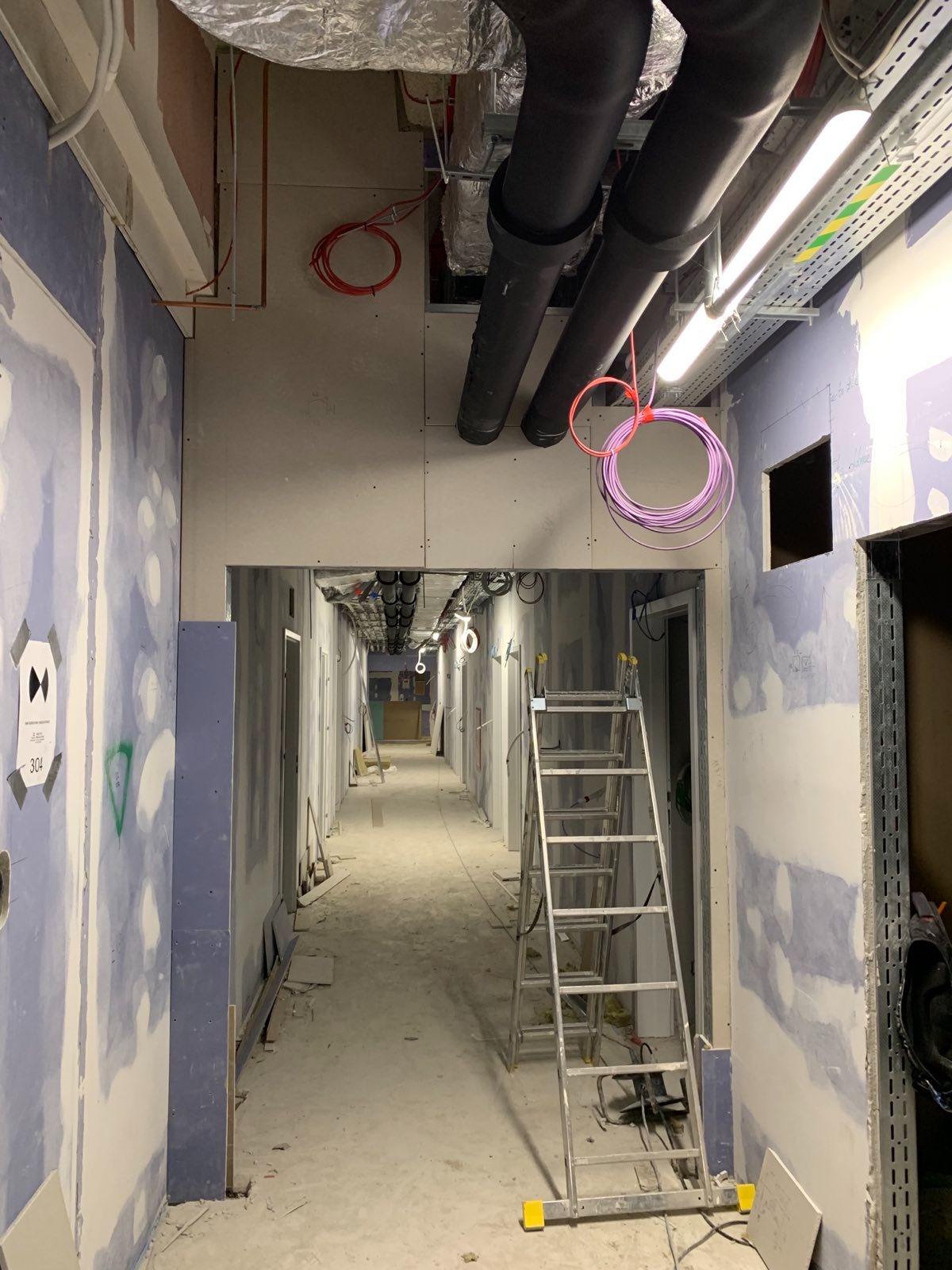 Korytarz w trakcie prac wykończeniowych: ściany z płyt gipsowo-kartonowych, widoczne instalacje pod sufitem, drabina. Charakterystyczne fioletowe kable zwisające z sufitu.