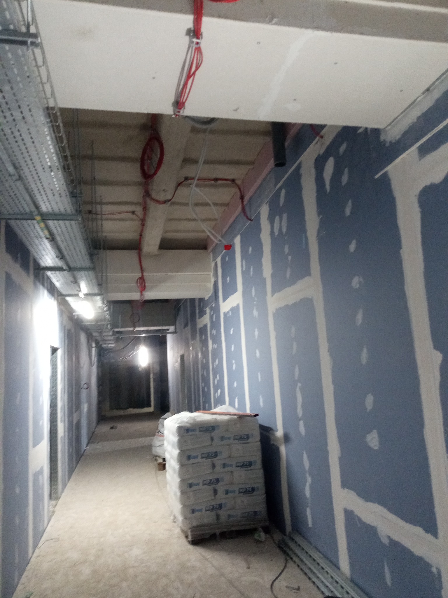 Korytarz w trakcie montażu płyt gipsowo-kartonowych, widoczne kable i profile. Na podłodze paleta z workami gładzi. Ściany w kolorze szarym, częściowo wyszpachlowane.