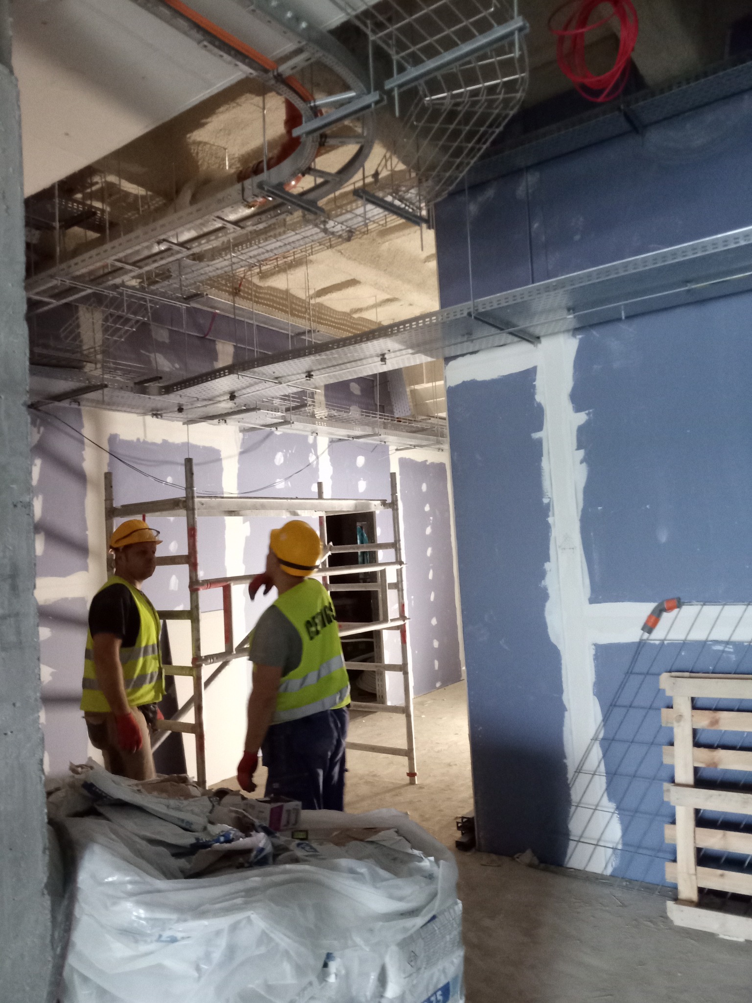 Montaż płyt gipsowo-kartonowych w biurze: pracownicy w kaskach i kamizelkach odblaskowych, rusztowanie, instalacje pod sufitem, ściany przygotowane do szpachlowania.