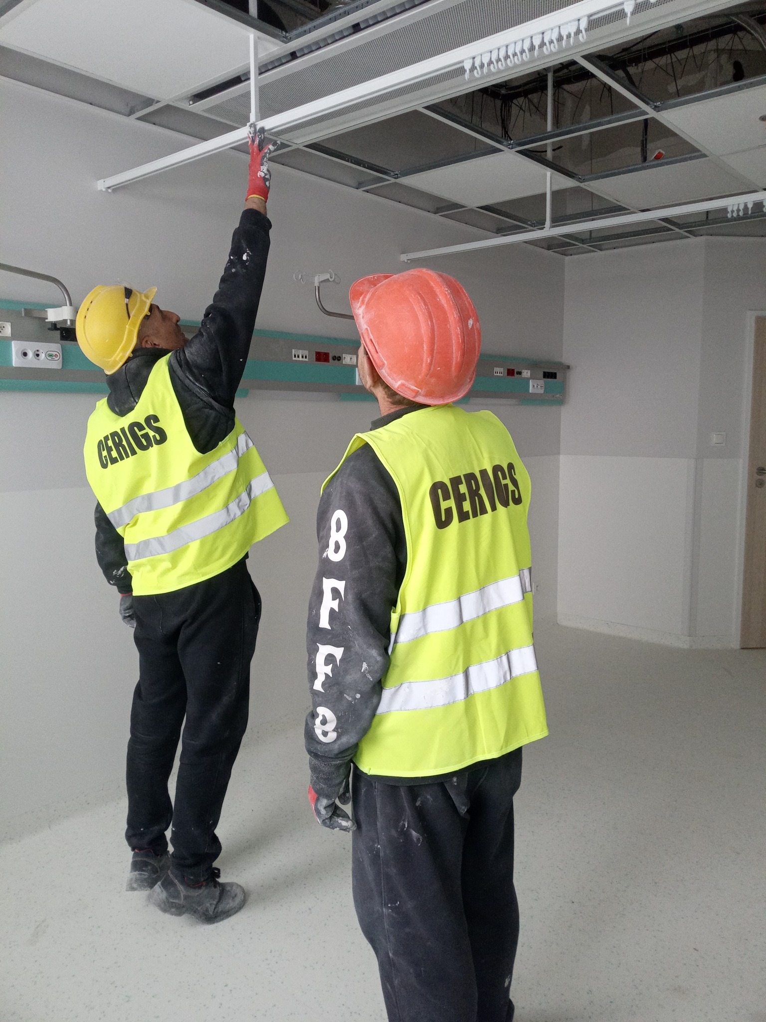 Dwóch monterów w kaskach i kamizelkach odblaskowych instaluje sufit podwieszany w pomieszczeniu z widocznymi elementami instalacji elektrycznej i wentylacyjnej.