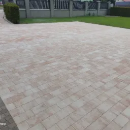 Nowo ułożona, beżowo-różowa kostka brukowa na podjeździe, z obrzeżem z ciemniejszej kostki, otoczona zielonym trawnikiem i tujami, widok z góry.