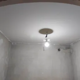 Pokój w trakcie przygotowania do malowania: surowe ściany z widocznymi śladami szpachlowania, świeżo pomalowany sufit z żarówką na kablu.