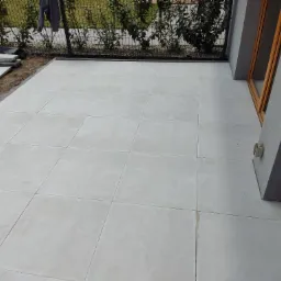 Jasne, kwadratowe płytki na zewnątrz budynku, z widocznymi zapasowymi płytkami i butami roboczymi w rogu, obok okna z drewnianą ramą i roślinności za ogrodzeniem.