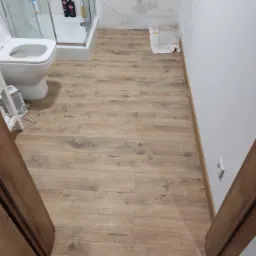 Ułożone panele podłogowe w łazience z widoczną kabiną prysznicową, toaletą i fragmentem grzejnika.