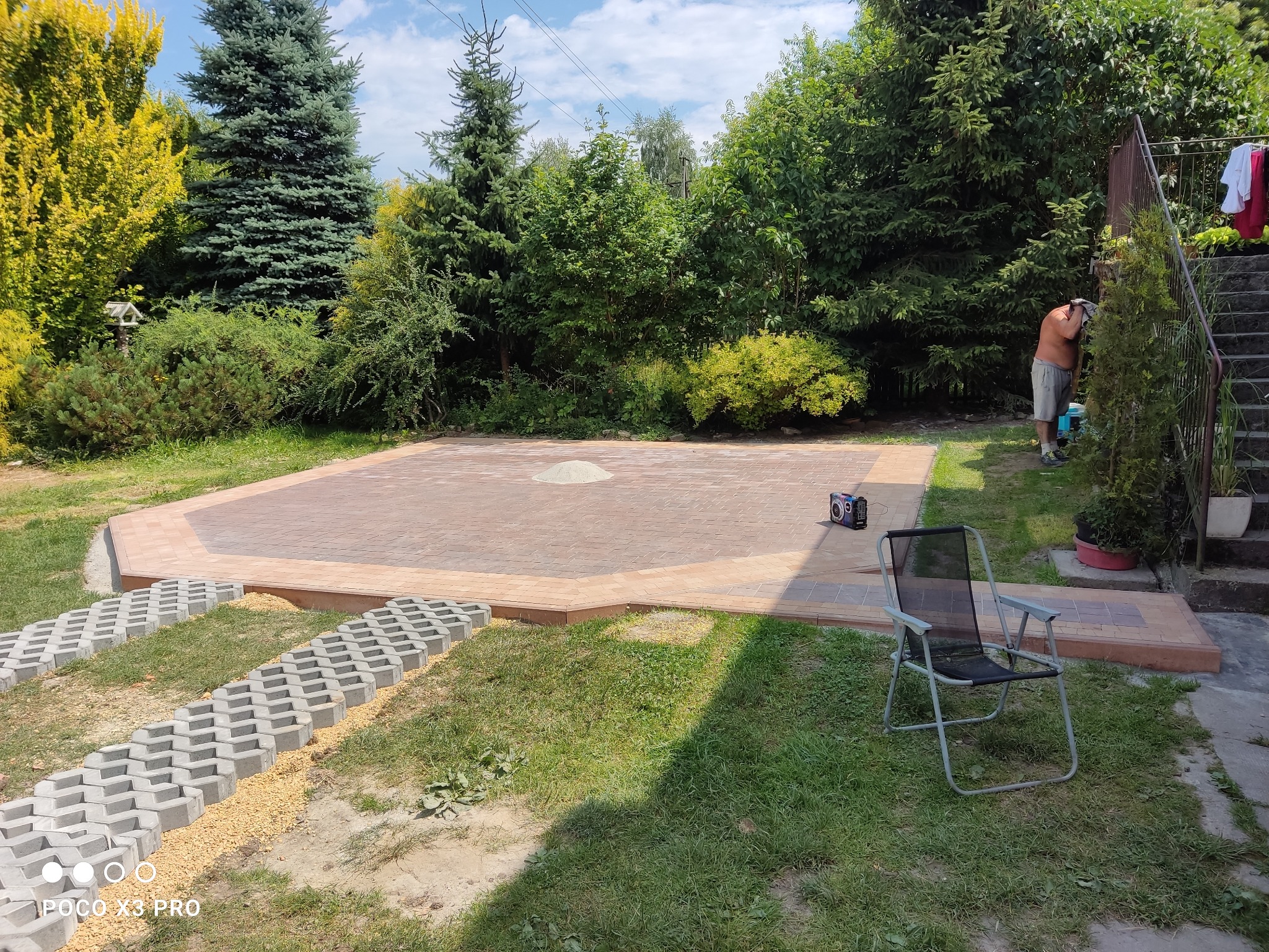 Ukończony taras z kostki brukowej w ogrodzie, widoczne ażurowe płyty betonowe z wypełnieniem żwirowym prowadzące do tarasu, radio na kostce, mężczyzna w tle.