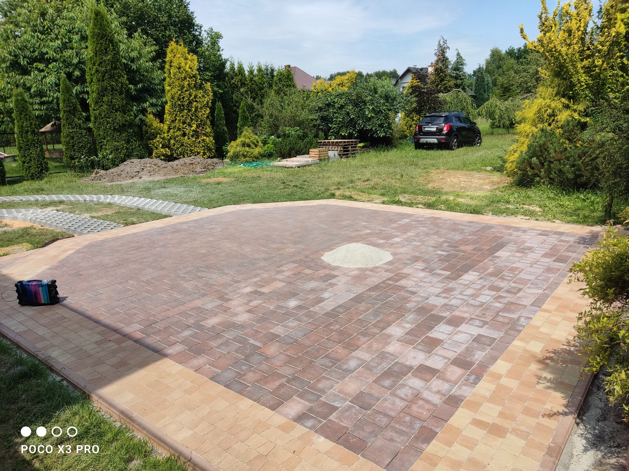 Rozległy plac z kostki brukowej w odcieniach brązu i beżu, z małą górką piasku pośrodku, otoczony zielenią i drzewami, z samochodem w tle.