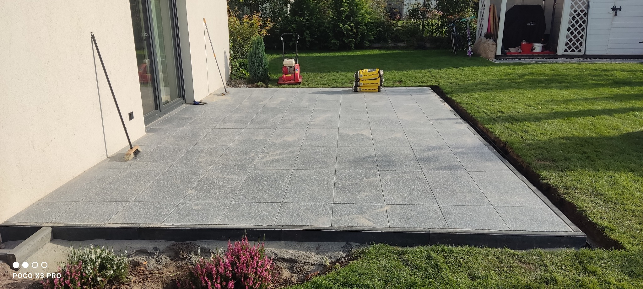 Ułożona szara kostka brukowa na tarasie, widoczna ubijarka i szczotka oparta o ścianę budynku, trawa i kwiaty w tle.