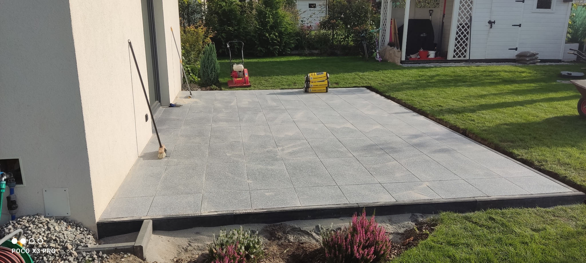 Ułożony taras z szarych płyt, z widocznym ubijakiem w tle, workami z piaskiem kwarcowym i częściowo wykończonym obrzeżem.