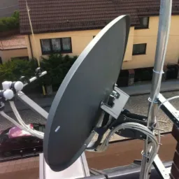 Szara antena satelitarna zamontowana na dachu budynku, widoczne kable i elementy mocujące, w tle dom z brązowym dachem i zaparkowany samochód.