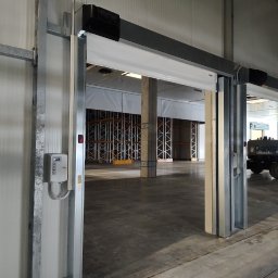 ProBram Adam Kurek - Otwarta automatyczna brama przemysłowa z szarą konstrukcją stalową i białym panelem, widok na puste wnętrze magazynu z regałami i betonową podłogą.