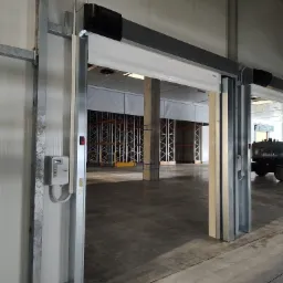 Otwarta automatyczna brama przemysłowa z szarą konstrukcją stalową i białym panelem, widok na puste wnętrze magazynu z regałami i betonową podłogą.