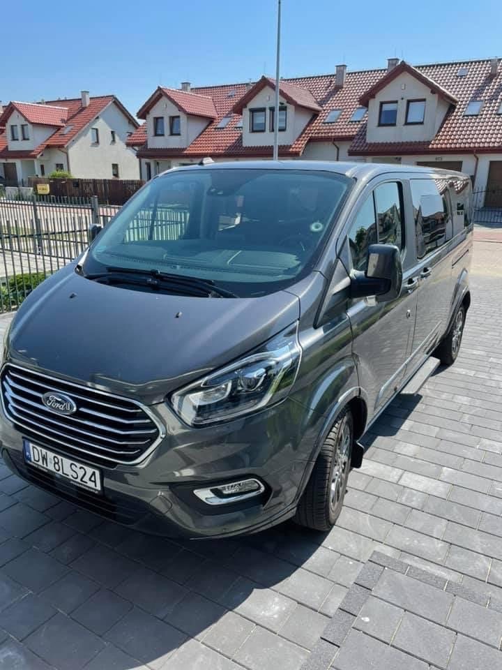 Szary Ford Transit Custom z polskimi tablicami rejestracyjnymi na tle osiedla domów jednorodzinnych.