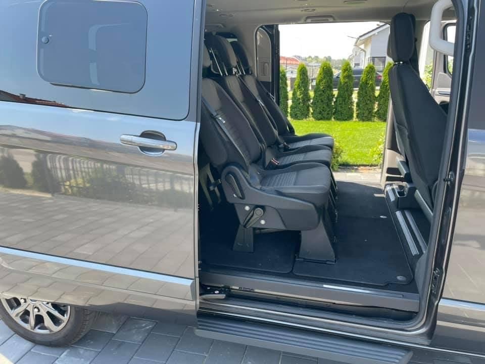 Wnętrze szarego busa z otwartymi drzwiami przesuwnymi, widoczne trzy rzędy czarnych foteli z zagłówkami i pasami bezpieczeństwa, za busem zadbany trawnik i tuje.