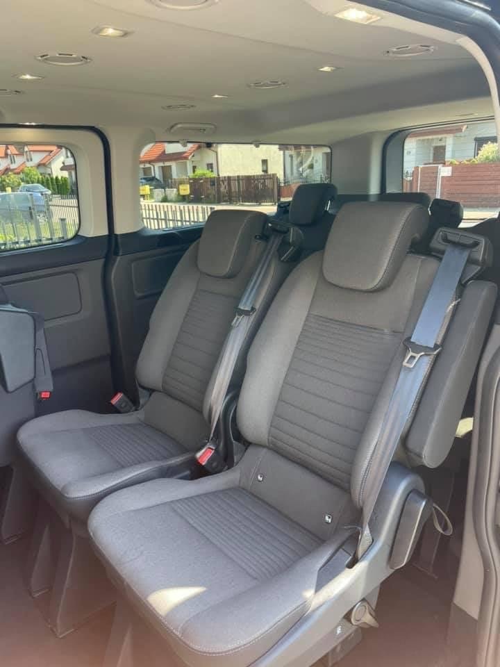 Wnętrze busa z dwoma rzędami foteli pasażerskich w szarej tapicerce, widok z boku na fotele z zapiętymi pasami bezpieczeństwa.