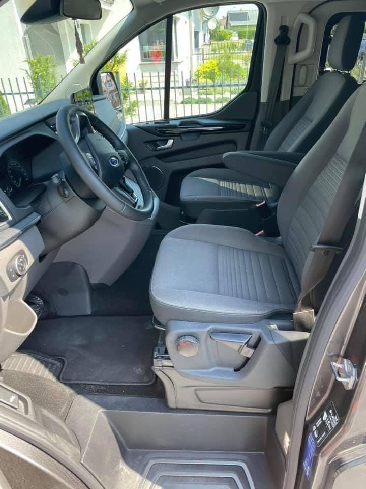 Wnętrze busa z szarymi fotelami, widok z zewnątrz przez otwarte drzwi, z regulacją fotela kierowcy i wiszącym odświeżaczem powietrza.