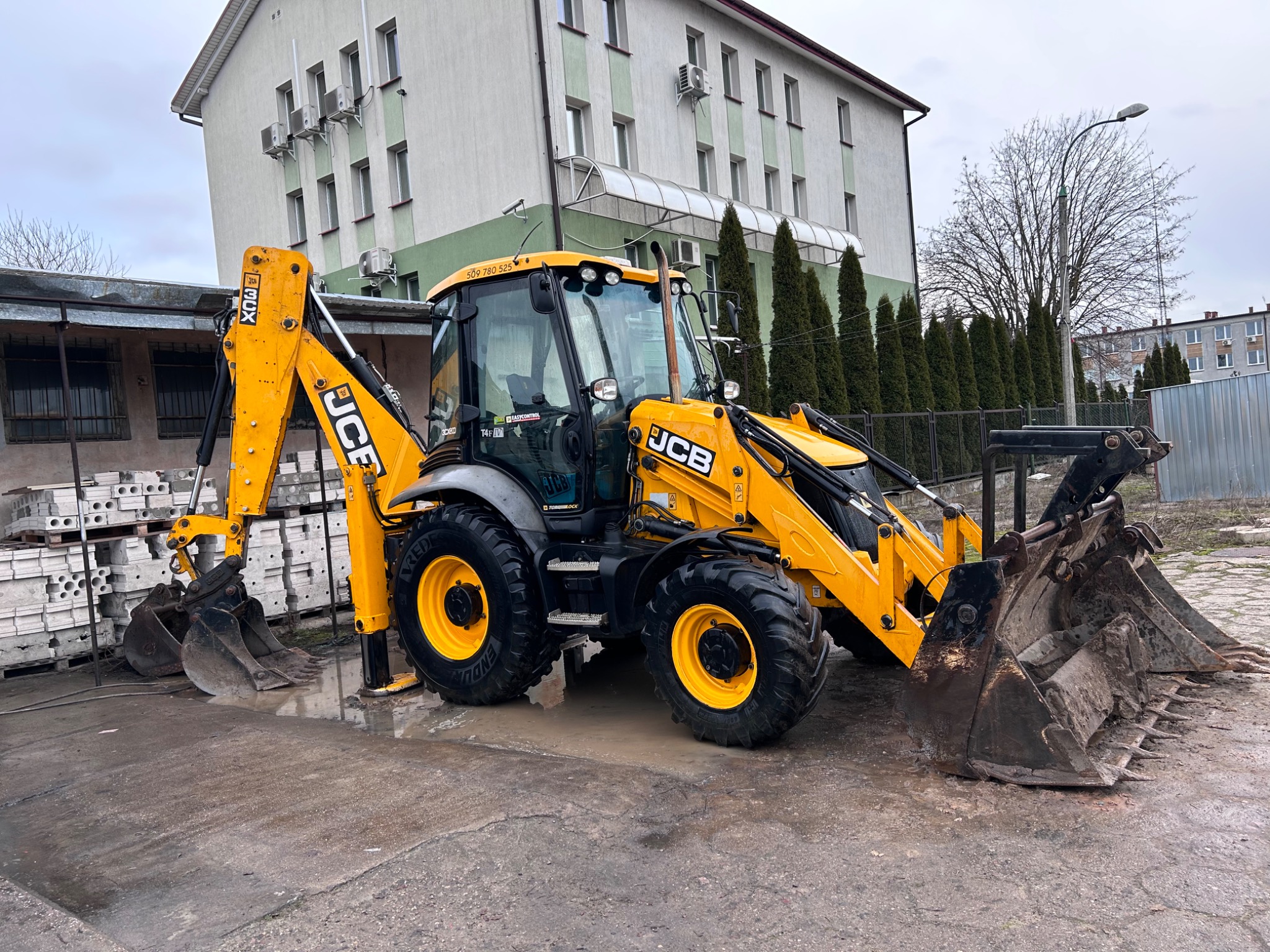 Żółto-czarna koparko-ładowarka JCB z dwoma łyżkami na mokrym placu budowy, budynek w tle.