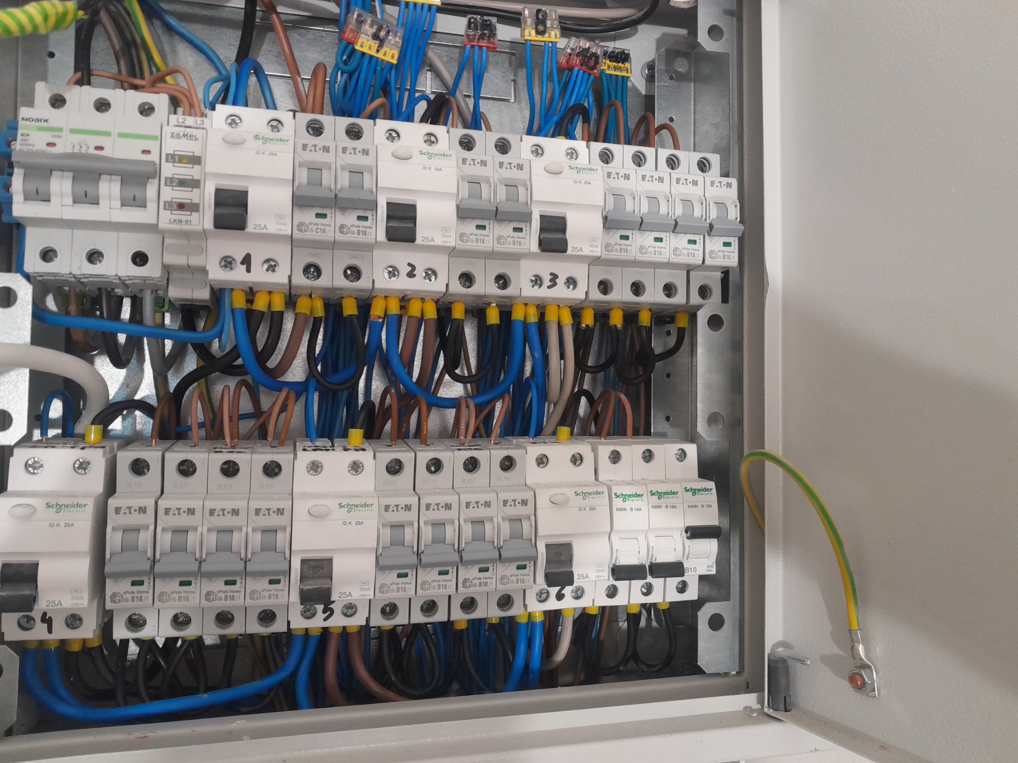Rozdzielnica elektryczna z zainstalowanymi bezpiecznikami różnych producentów (Schneider, Eaton, Noark, Zamel) i podłączonymi przewodami o różnych kolorach, widoczne oznaczenia prądowe i napięciowe...