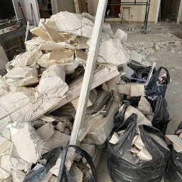 Wywóz Śmieci Gruzu Gabarytów - Sterta gruzu z wyburzeń w pomieszczeniu, częściowo spakowana w czarne worki, z widocznym fragmentem rusztowania i osobą w tle.