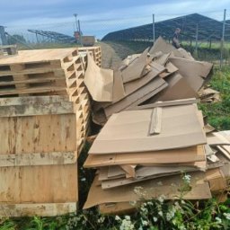 Wywóz Śmieci Gruzu Gabarytów - Stos drewnianych palet i sterta pociętej tektury na trawie, w tle panele słoneczne i osoba na drodze.