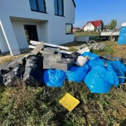 Sterta odpadów budowlanych w workach i luzem przed domem w Inowrocławiu, z widoczną przyczepą i przenośną toaletą w tle.