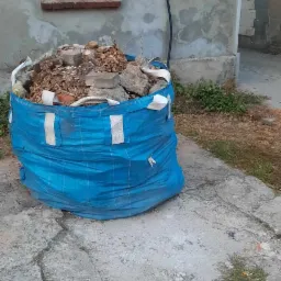 Niebieski worek typu Big Bag wypełniony gruzem budowlanym stoi na popękanym betonowym podłożu przed ceglanym budynkiem z białym oknem.