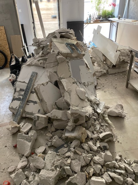 Stos gruzu i fragmentów ściany gipsowo-kartonowej z widocznym metalowym profilem na podłodze wewnątrz pomieszczenia, po zakończonych pracach demontażowych.