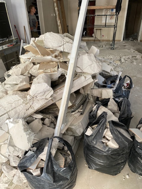 Sterta gruzu z wyburzeń w pomieszczeniu, częściowo spakowana w czarne worki, z widocznym fragmentem rusztowania i osobą w tle.