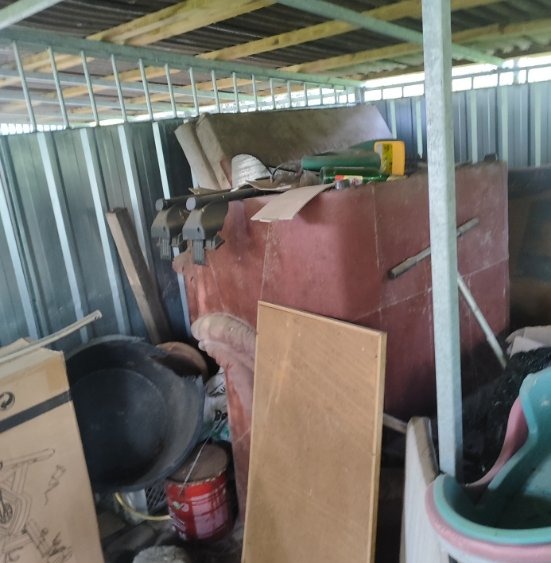 Zastawiony blaszany garaż z różnorodnymi przedmiotami, w tym meblami, narzędziami i pojemnikami, pod metalowym zadaszeniem.