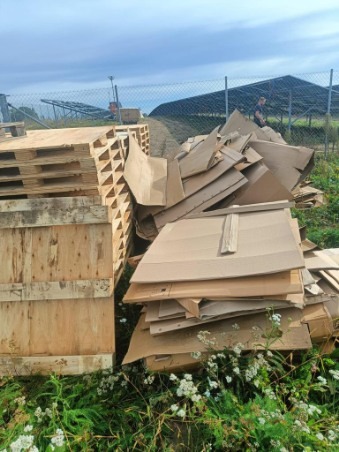 Stos drewnianych palet i sterta pociętej tektury na trawie, w tle panele słoneczne i osoba na drodze.