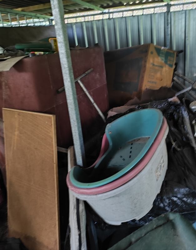 Stare, plastikowe wanienki dziecięce w stercie nieposegregowanych przedmiotów, w tym kartonów i elementów mebli, składowanych w zadaszonym pomieszczeniu.