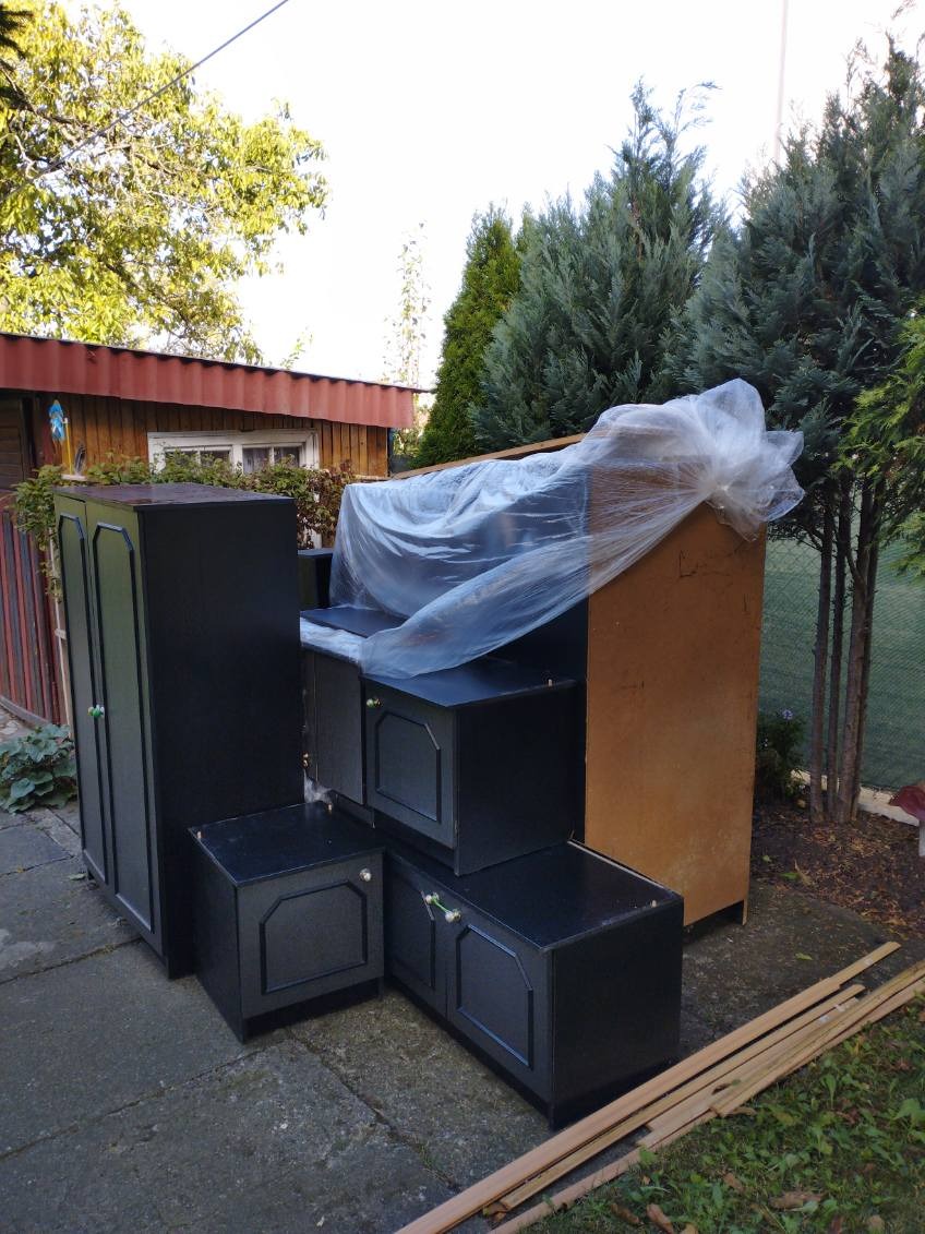 Częściowo zabezpieczone folią meble, prawdopodobnie do wyniesienia, ustawione na zewnątrz na betonowej nawierzchni, w tle widać iglaki i drewniany budynek.