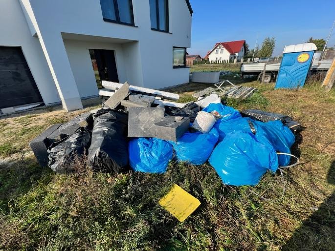 Sterta odpadów budowlanych w workach i luzem przed domem w Inowrocławiu, z widoczną przyczepą i przenośną toaletą w tle.