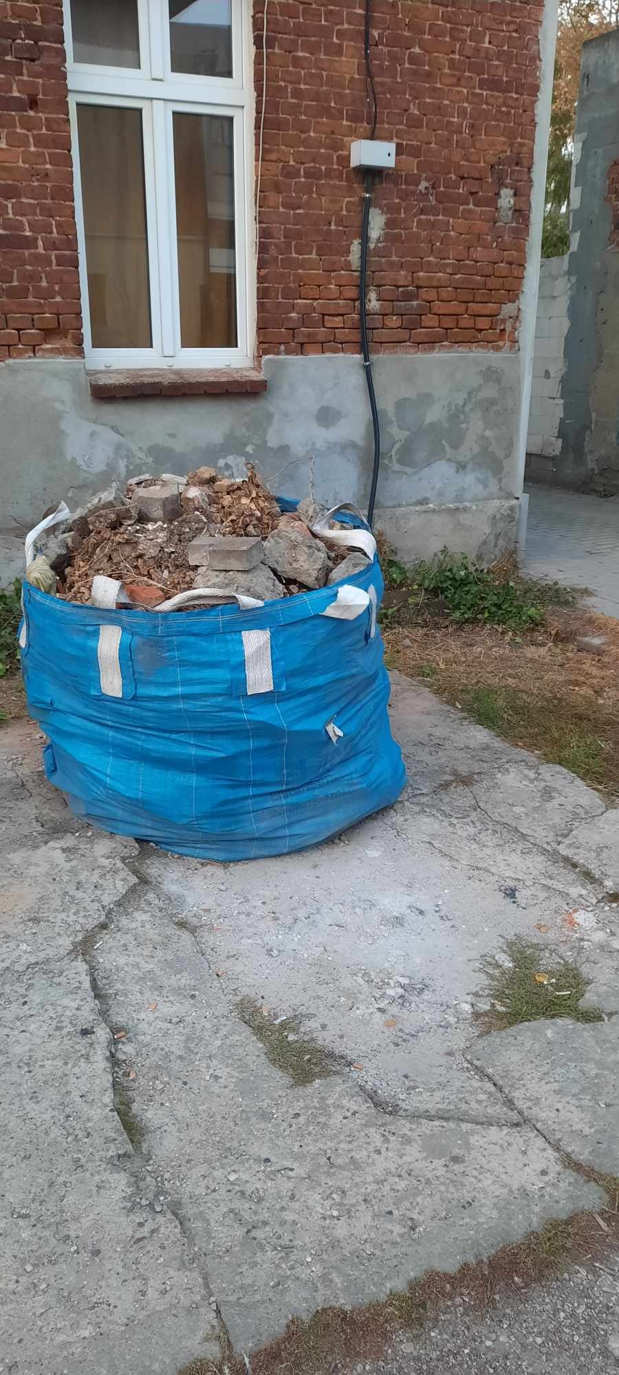 Niebieski worek typu Big Bag wypełniony gruzem budowlanym stoi na popękanym betonowym podłożu przed ceglanym budynkiem z białym oknem.
