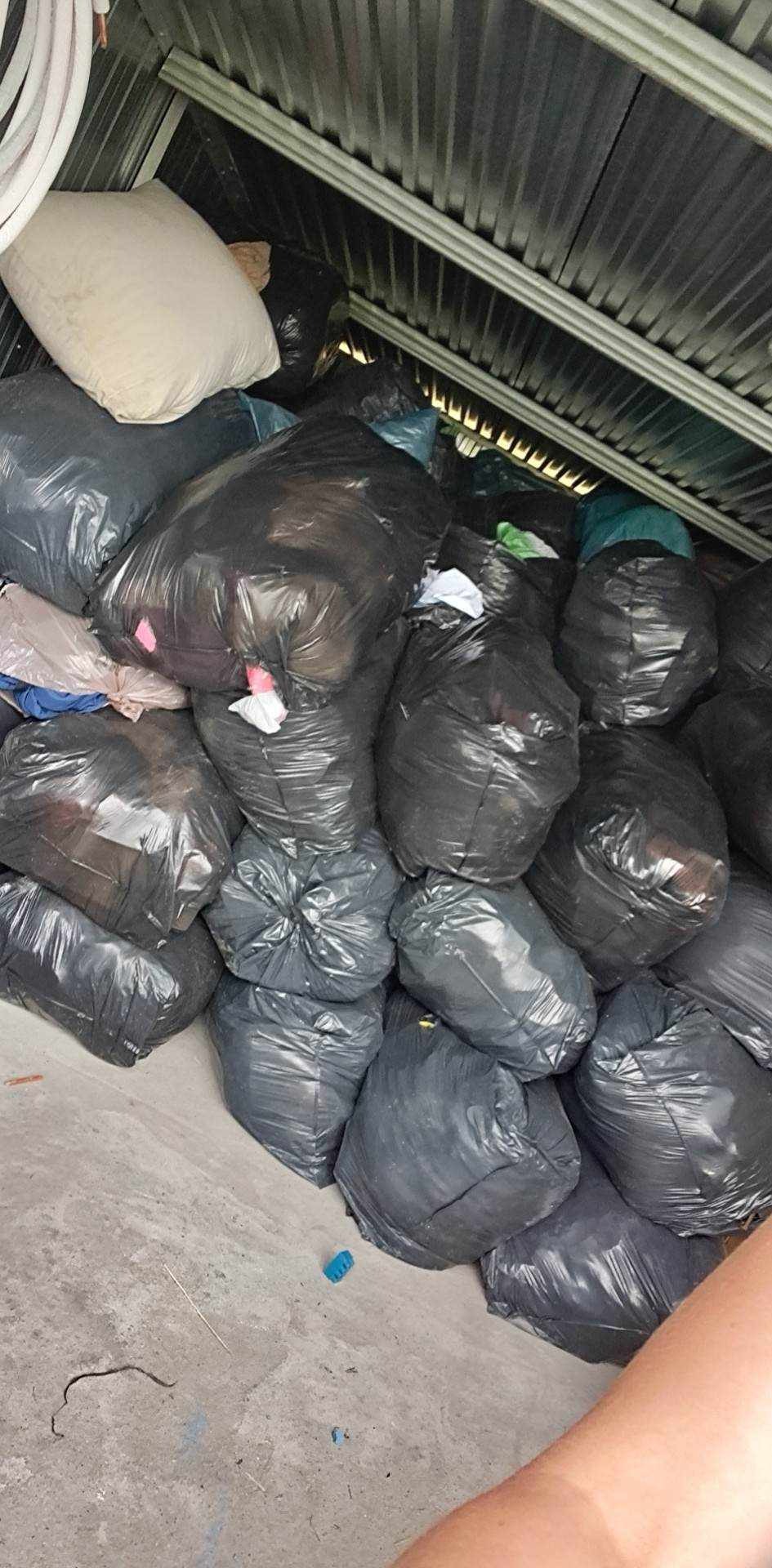 Stos czarnych worków na śmieci, częściowo wypełnionych, w pomieszczeniu o metalowym suficie, jeden biały worek na górze.