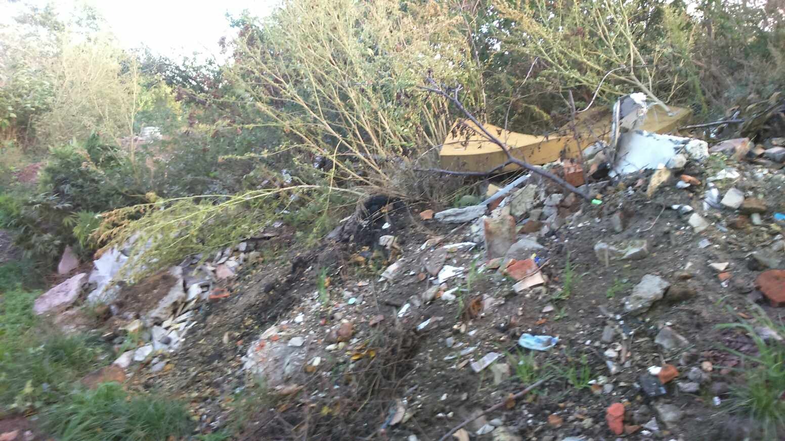 Sterta gruzu zmieszanego z ziemią i roślinnością, fragmenty cegieł i betonu, w tle krzaki i mały fragment żółtego elementu.