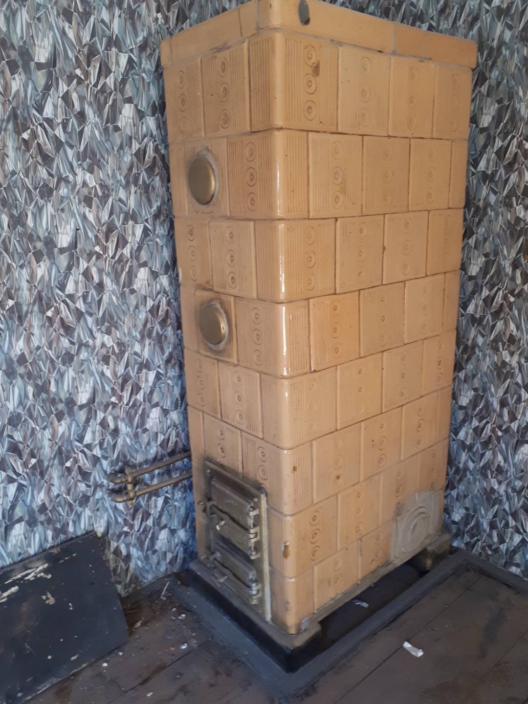 Stary piec kaflowy w odcieniach beżu z okrągłymi zdobieniami, widoczne drzwiczki i metalowe elementy, na tle ściany z wzorzystą tapetą, na podłodze widoczne ślady użytkowania.