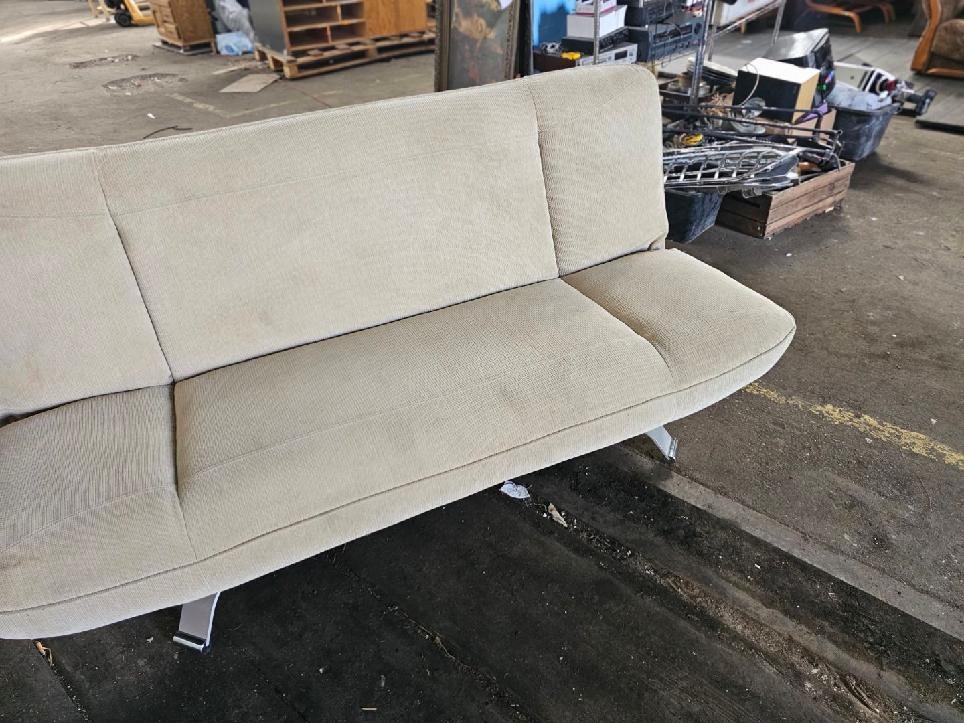 Beżowa sofa z trzema siedziskami i metalowymi nogami, ustawiona w hali z widocznym bałaganem i różnymi przedmiotami w tle.