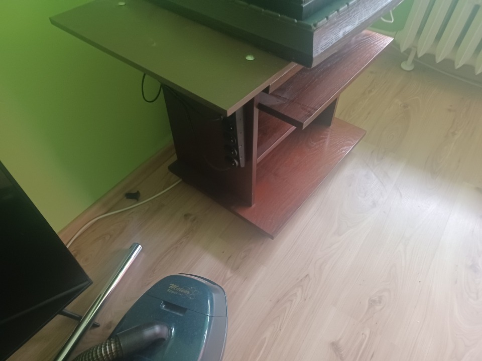 Stary odkurzacz Meteor Super na drewnianej podłodze obok stolika RTV z zieloną ścianą w tle.