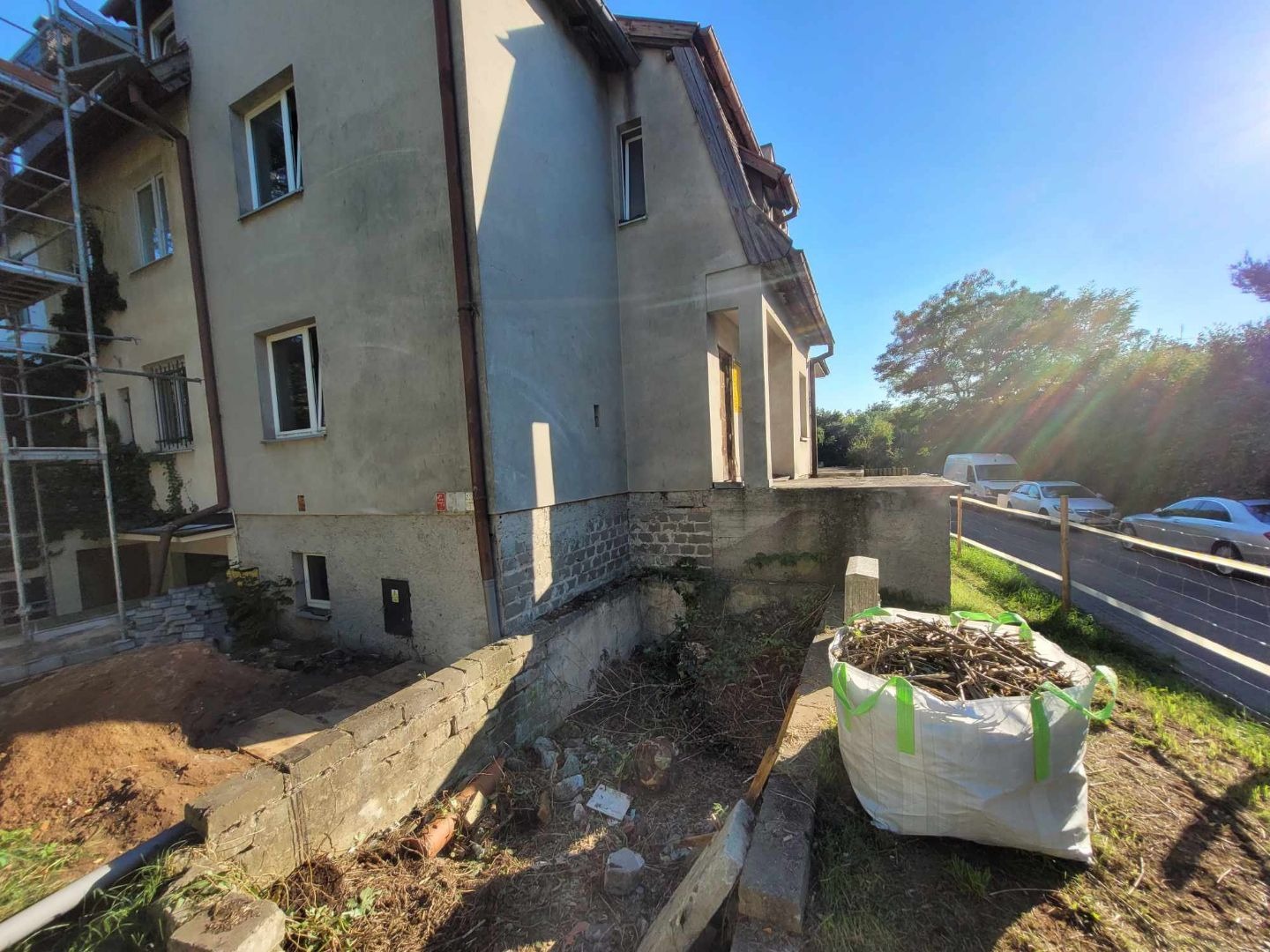 Przygotowania do rozbiórki: widok na budynek z rusztowaniem, odsłonięty fundament i duży worek z gałęziami na pierwszym planie w słoneczny dzień.