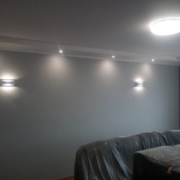 krzysztof kaleński - Świeżo pomalowana szara ściana z zamontowanymi kinkietami i oświetleniem punktowym w suficie, meble zabezpieczone folią ochronną.